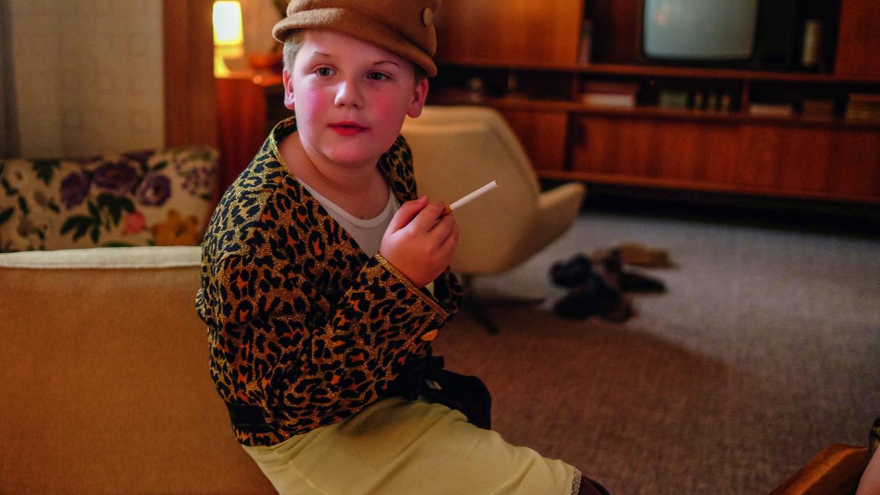 Das Kind Hans Peter als Frau verkleidet mit Hut, Leopardenmustermantel und Zigarette in der Hand sitzt in einem Wohnzimmer der 1970er-Jahre.