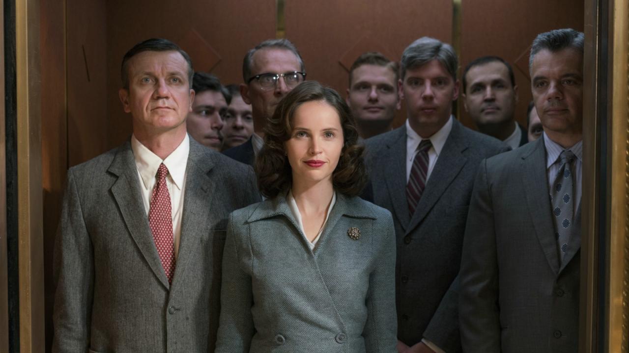 Still aus dem Biopic DIE BERUFUNG: Eine junge Frau, gekleidet in einem grau-blauem Kostüm im Stil der 1950er, steht in einem Fahrstuhl. Sie ist umgeben von Männern in grauen Anzügen.
