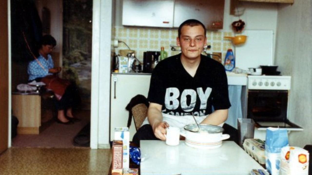 Szenebild aus dem Dokumentarfilm STAU - JETZT GEHT'S LOS: Ein junger Mann sitzt an einem Tisch in der Küche. Auf seinem T-Shirt steht "Boy". Im Hintergrund sitzt die Mutter in einem anderen Zimmer.
