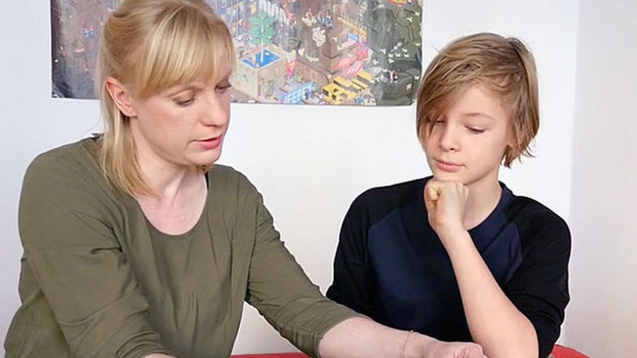 Videotutorial (Szenenbild): Einer blonde Frau und ein blonder Junge sitzen an einen Tischen und schauen runter.