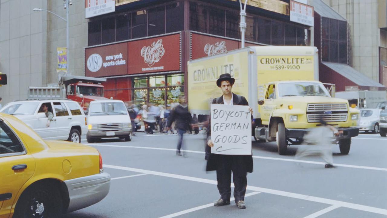Schlingesief – In das Schweigen hineinschreien, Szenenbild: Ein Mann steht mit einem Protestschild auf einer belebten Straßenkreuzung in New York