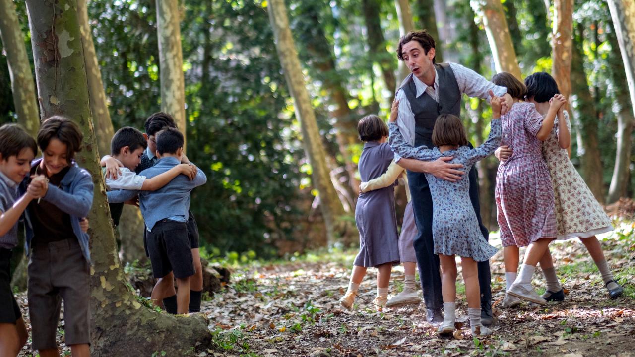 ilmbild aus dem Drama DER LEHRER, DER UNS DAS MEER VERSPRACH: Ein Mann und mehrere Kinder, alle im Stil der 1940er gekleidet, tanzen paarweise zwischen Bäumen.