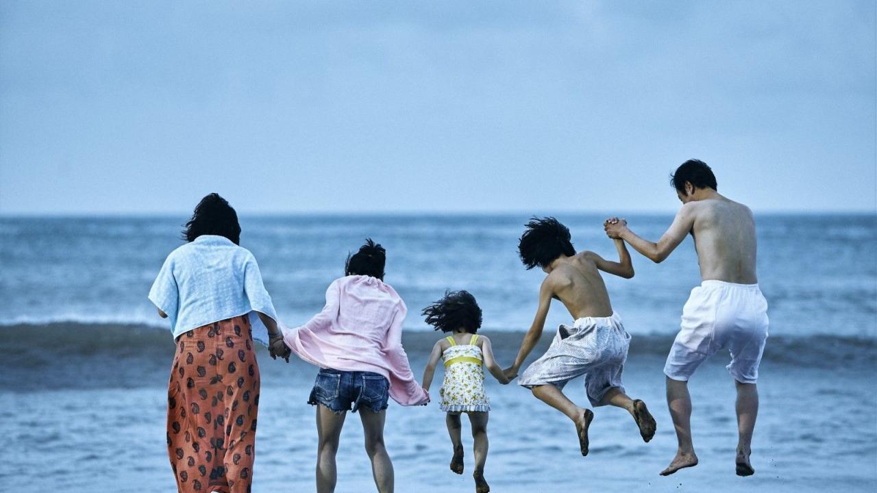Shoplifters, Szenenbild: Rückenansicht von zwei Frauen, einem kleinen Mädchen, einem Jungen und einem Mann am Meer. Sie halten sich an den Hängen und springen in die Luft.