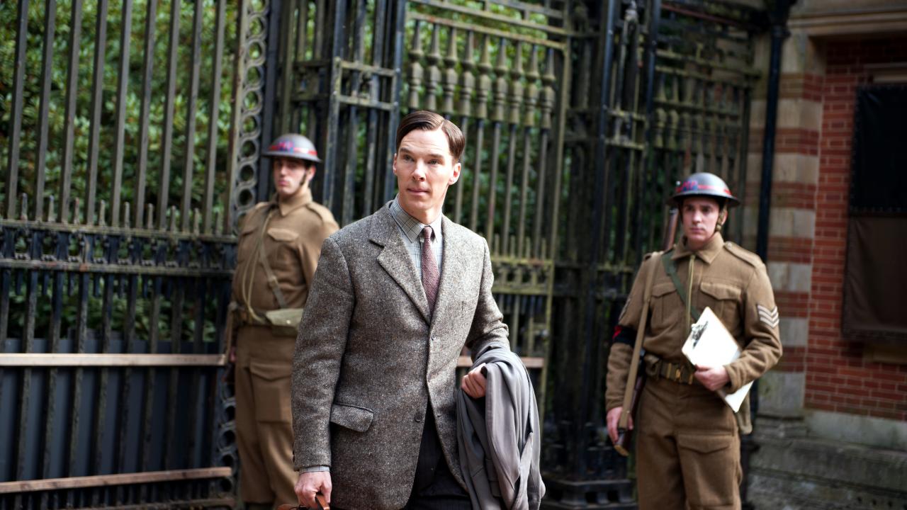 Filmstill aus THE IMITATION GAME: Alan Turing (Benedict Cumberbatch) geht sich vor einem großen, gußeisernen Tor. Hinter ihm stehen britische Soldaten,