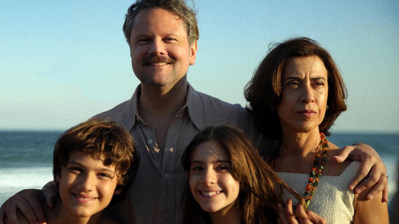Still aus dem Drama FÜR IMMER HIER: Ein Familienfoto am Meer in warmen Farbe: Vater und Mutter stehen hinter den Kindern. Alle lächeln, außer die Frau.