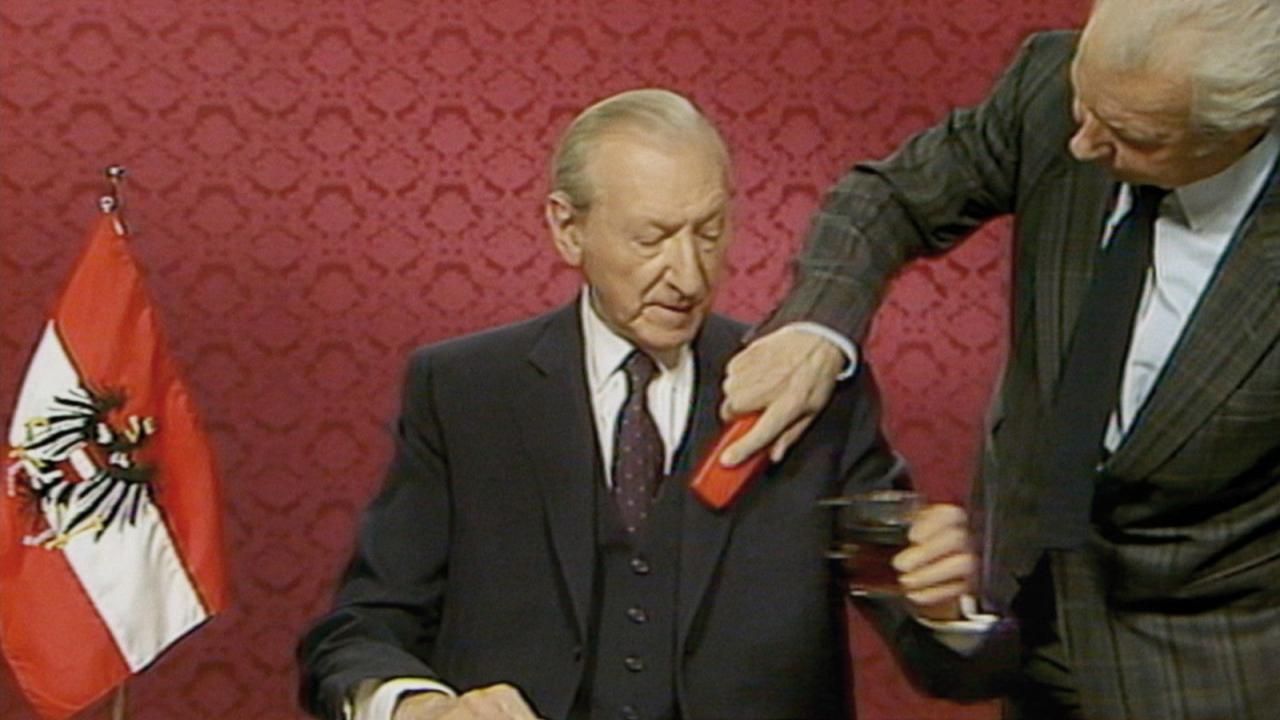 Waldheims Walzer, Szenenbild: Der österreichische Politiker Kurt Waldheim kurz vor eine Rede im Fernsehen. Jemand bürstet ihm die Jacke ab. Er hält ein Glas in seiner Hand. 