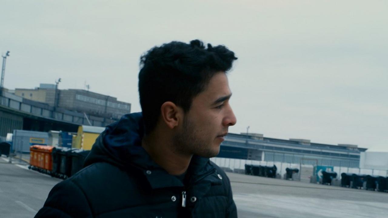 Zentralflugafen THF, Szenenbild: Ein junger Mann mit dunklen Haaren und blauen Anorak im Profil, im Hinterrgund ist das Gebäude des Berliner Flughafens Tempelhof.