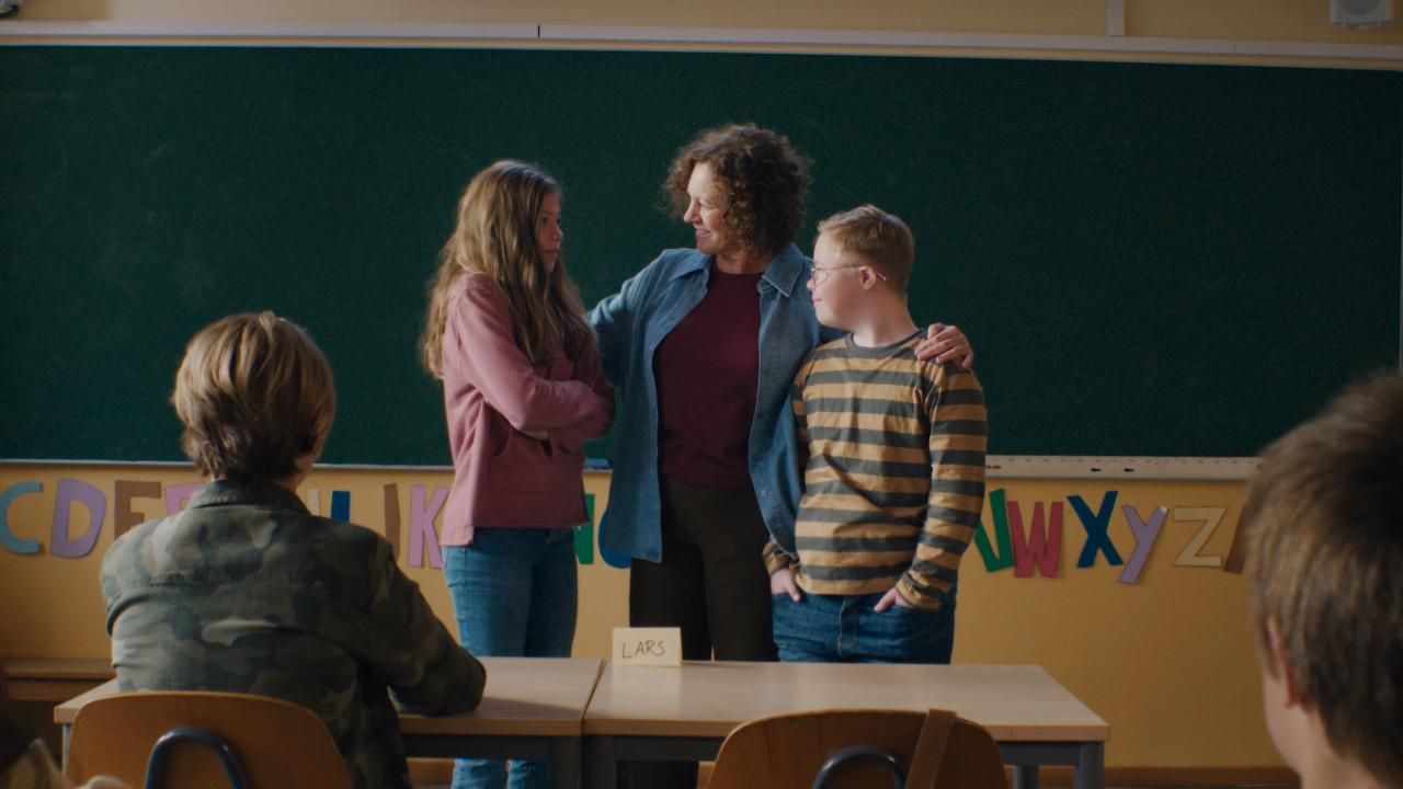Filmstill aus LARS IST LOL: Vor einer Schultafel steht eine Frau, die ein Mädchen und einen Jungen miteinander bekannt macht. Das Mädchen hat die Arme verschränkt, der Junge lächelt sie an. Er hat das Down-Syndrom.