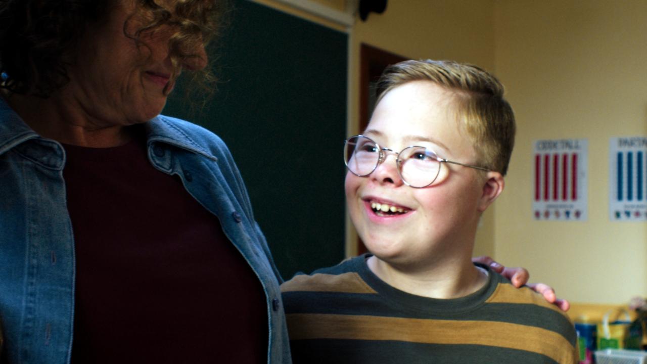 Filmstill aus LARS IST LOL: Ein Junge mit Down-Syndrom schaut lachend eine Frau an, die ihren Arm um ihn gelegt hat. Sie stehen in einem Klassenzimmer.