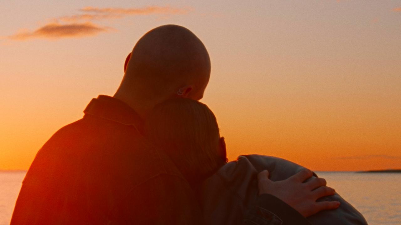 Still aus dem Drama WENN DAS LICHT ZERBRICHT: Im Gegenlicht sitzen ein junger Mann und eine junge Frau am Meer und schauen auf dasLicht der untergehenden Sonne. Der Mann hat den Arm un die Frau gelegt, die sich an ihn anschmiegt. Man sieht beide in der Rückenansicht.