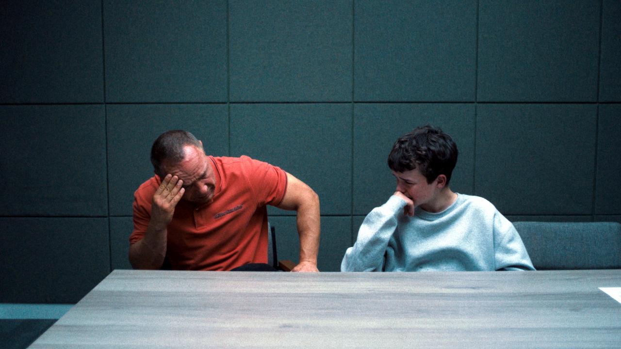 Filmstill aus der Mini-Serie ADOLESCENCE: Ein Mann in rotem Poloshirt und ein Junge im weißen Sweatshirt sitzen in einem Verhörraum nebeneinander. Der Mann stützt seinen Kopf in die Hand und schaut nach unten. Der Junge hat seine Hand vor dem Mund und und blickt zu dem Mann.