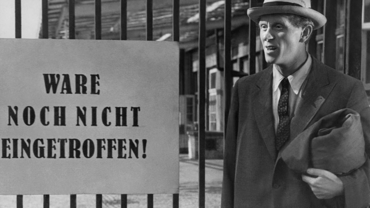 Szenenbild aus dem Film BERLINER BALLADE (1948): Ein Mann in einem zu weiten Anzug steht vor einem Gittertor mit einem Schild. Darauf steht "Ware noch nicht eingetroffen".