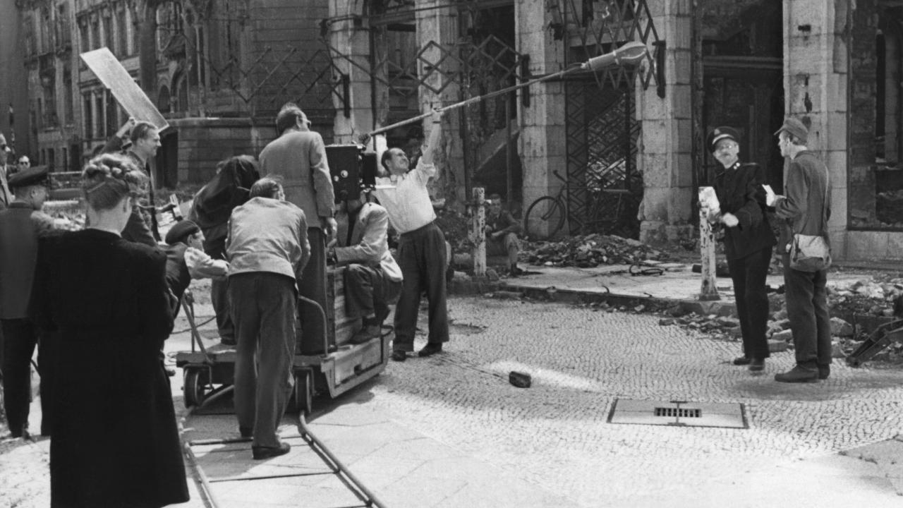 Setfoto vom Film BERLINER BALLADE (1948): Vor einer Trümmerkulisse stehen links ein Filmteam mit einem Kamerawagen auf Schienen und rechts zwei Darsteller. Ein Mitarbeiter hält eine Mikrofonangel in ihre Richtung.
