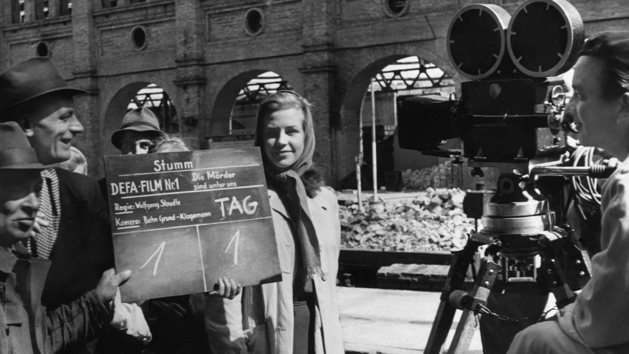 Setaufnahme vom Film DIE MÖRDER SIND UNTER UNS (1946): Die junge Schauspielerin Hildegard Knef steht neben weitere Personen hinter eine Filmklappe und einer Kamera. Hinter ihr eine Häuserruine zu sehen.