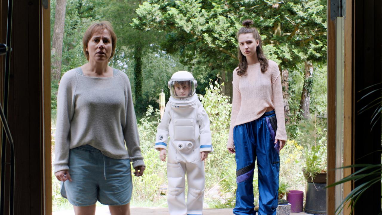 Still aus dem Kinderfilm GRÜSSE VOM MARS: Eine Frau, ein Junge und ein Mädchen im Teenageralte stehen vor einer geöffnetten Terrassentür und schauen überrascht in die das Zimmer. Der Junge trägt einen Astronautenanzug.