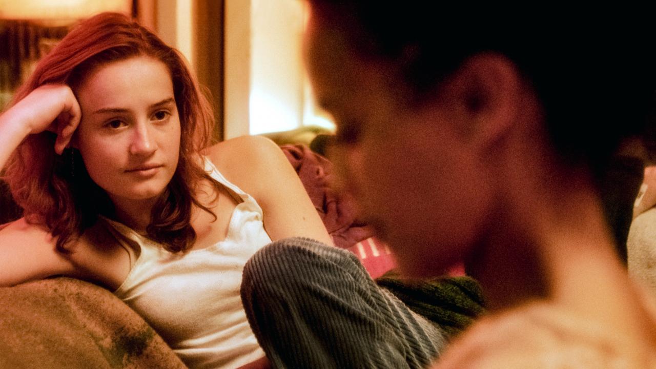 Still aus dem Film OSLO-STORIES: TRÄUME: Eine junge Frau sitzt auf einem Sofa und schaut eine andere Frau an, die im Vordergrund des Bilds unscharf zu erkennen ist. Das Bild ist in warmen Farbtönen gehalten.