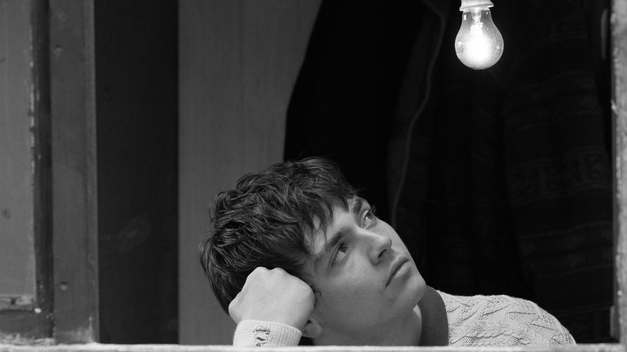 Schwarz-weißes Filmstill aus dem Drama YURT: Eine Junge sitzt an einem geöffneten Fenster und hat seinen Kopf auf eine Hand gestützt. Er schaut schräg nach oben zu einer nackten Glühbirne. 