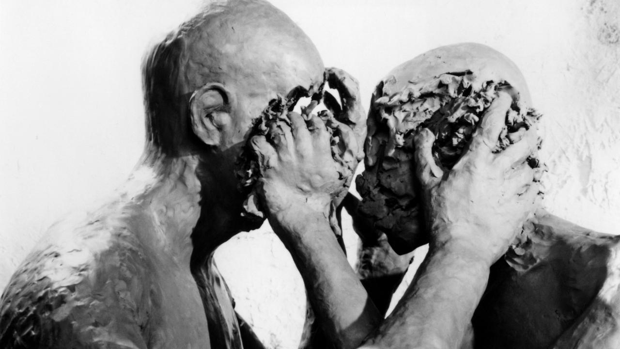 Filmstill aus dem Experimentalfilm TÜCKEN DES GESPRÄCHS: Zwei glatzköpfige Menschen im Profil halten sich an den Köpfen fest. Ihre Gesichter sind verschwunden wirken wie geknetet.