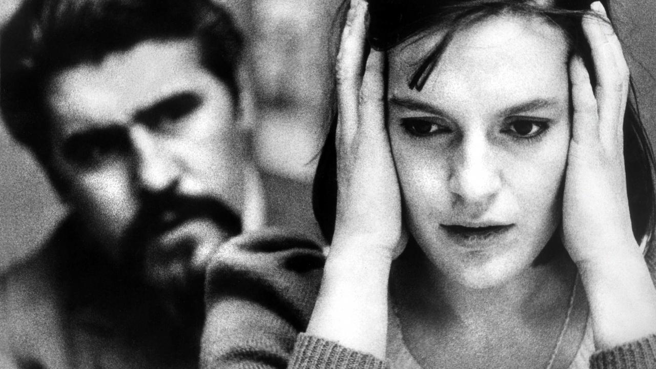 Schwarz-weißes Stil aus dem Drama DIE VERLORENE EHRE DER KATHARINA BLUM: Eine junge Frau in Nahuafnahme steht im Vordergrund des Bilds. Sie hält beide Hände seitlich an ihr Gesicht. Hinter ihr ist unscharf ein Mann zu erkennen, der in ihre Richtung schaut.