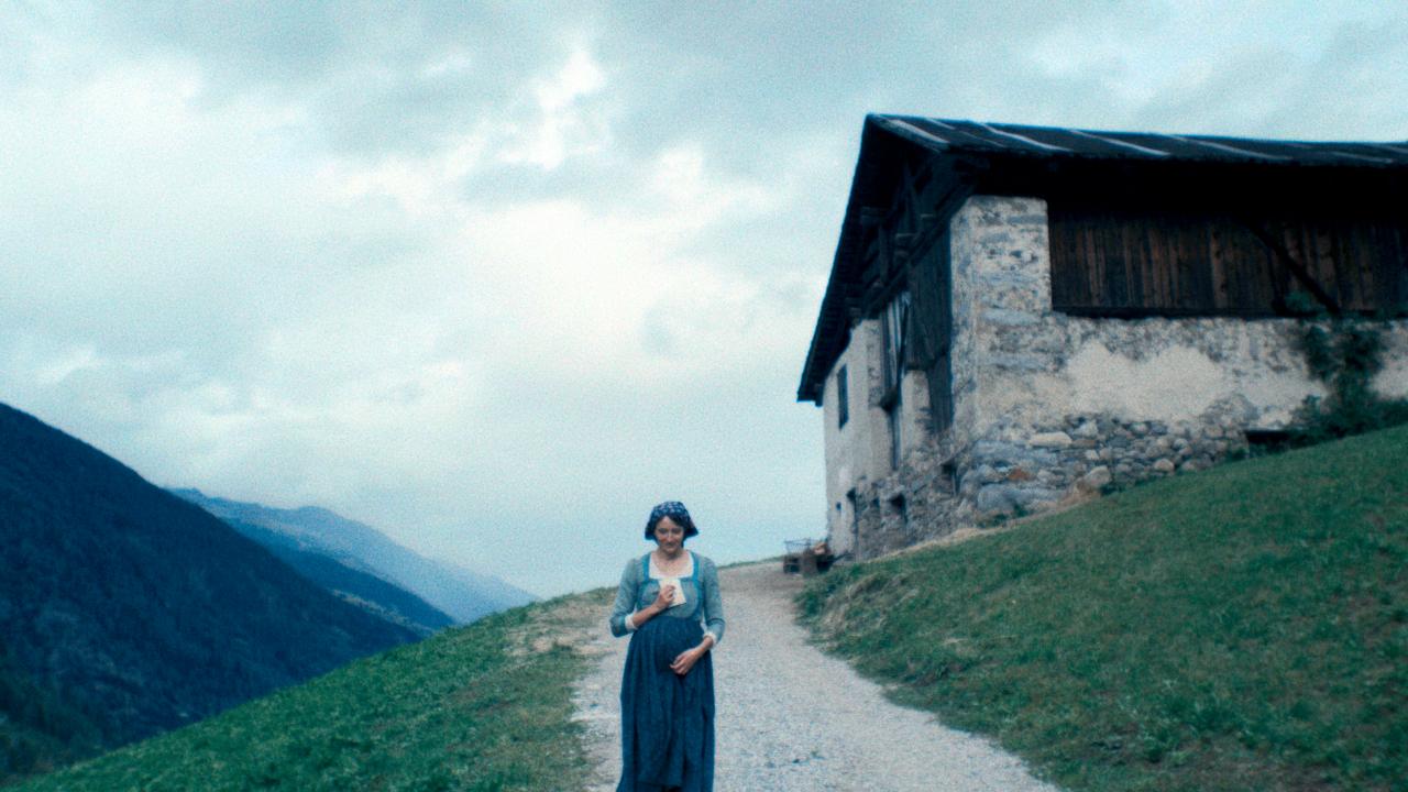 Filmstill aus dem Drama VERMIGLIO: In einer Alpenlandschschaft geht eine junge, schwangere Frau einen Weg entlang. Sie ist in bildmittig angeordnet. Neben ihr steht ein steineren Haus.