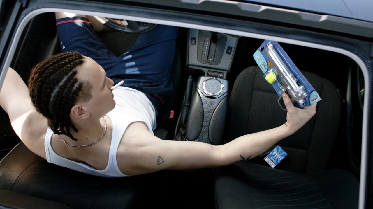Still aus dem Drama EINER VON UNS: Aus der Aufsicht: Durch das geöffnete Schiebedach eines Autos sieht man einen jungen Mann am Steuer, der sich eine Pistole in einer Verpackung anschaut.