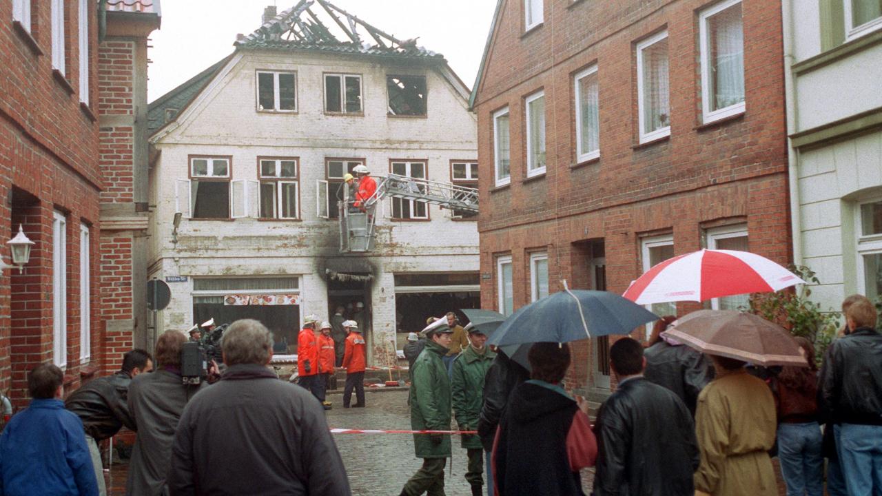 Foto des Wohnhauses Mühlenstraße 9 in Mölln nach dem Brandanschlag von 23. November 1992: Eingerahmt von den Häuser der auf sie zuführenden ist Nebenstraße ist die Fassade eine offensichtlich durch einen Brand zerstörten Hauses mit verkohltem Dachstuhl und um Fenster und Tür herum verrusten Altbaus zusehen. Vor dem Gebäude stehen Feuerwehrleute. Ein Absperrband trennen Polizisten von einen Gruppe von Zivilist/-innen die zum Haus schauen.