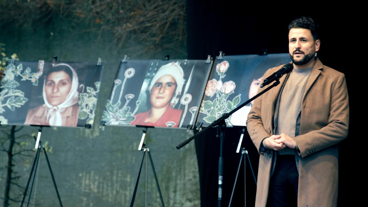 Still aus dem Dokumentarfilm DIE MÖLLNER BRIEFE:  İbrahim Arslan steht auf einer Bühne vor einem Mikrofon. Neben ihm stehen Bilder seiner verstorbenen Angehörigen. 