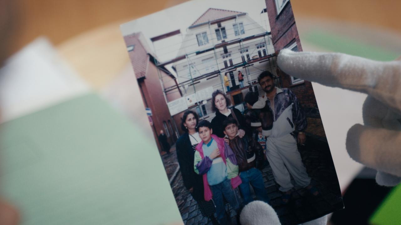 Still aus dem Dokumentarfilm DIE MÖLLNER BRIEFE: Bild einer Fotografie, auf der eine türkische Familie zu sehen ist, die vor dem Haus in Mölln steht, auf das 1992 ein rassistischer Brandanschlag verübt worden ist.