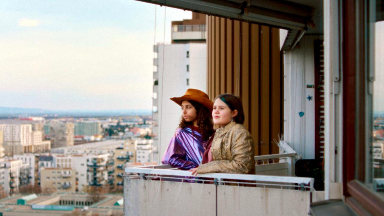 Still ais dem Drama WENN DU ANGST HAST NIMMST DU DEIN HERZ IN DEN MUND UND LÄCHELST: Zwei Mädchen im Teenageralter stehen in einer Hochhaissiedlung auf einem Balkon.