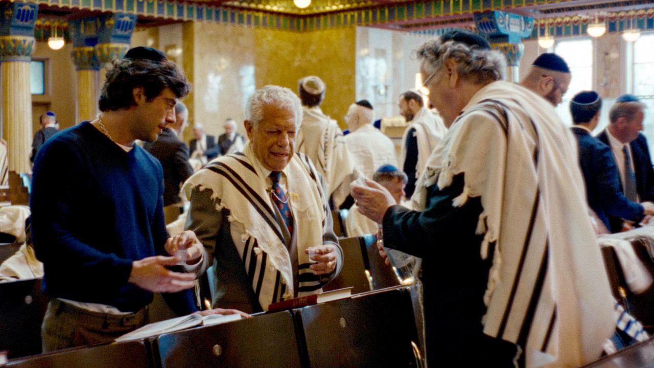 Still aus der Serie DIE ZWEIFLERS: Ein junger Mann mit Kippa und zwei ältere herren mit Gebetsschal stehen nebeneinander in einer Synagoge.