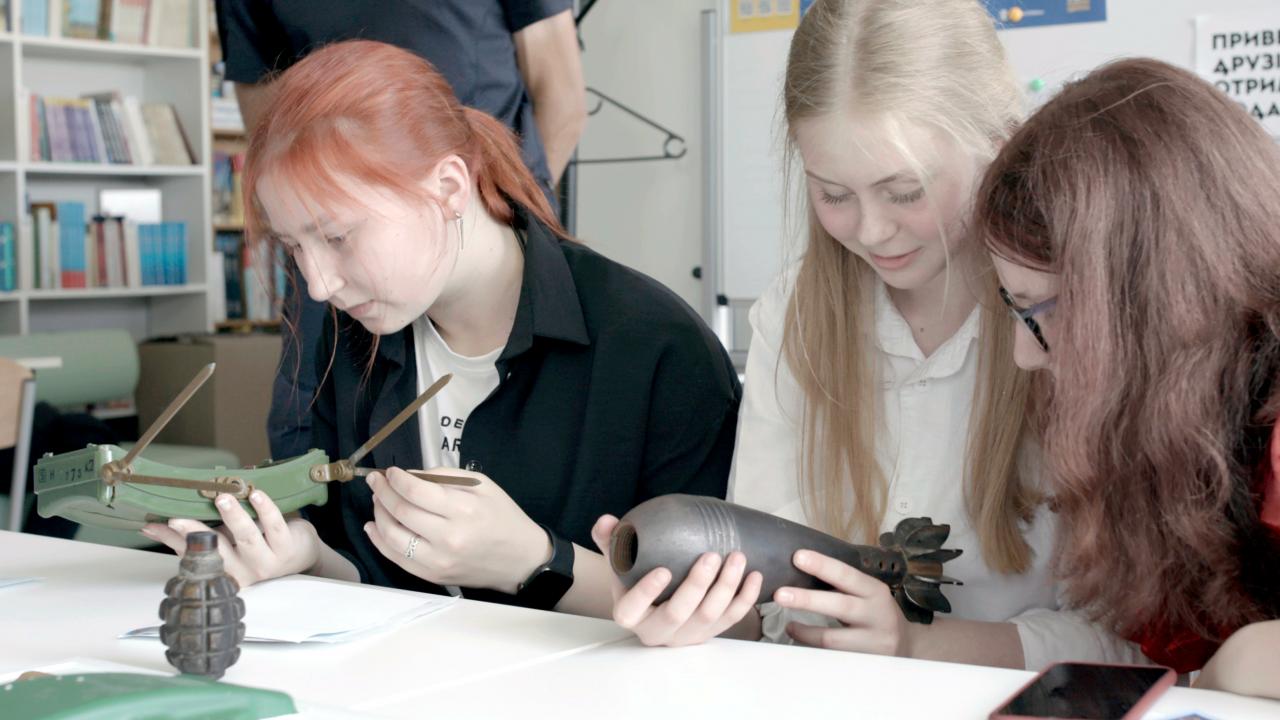 Still aus dem Dokumentarfilm SPLITTER AUS LICHT: Drei Teenagerinnen sitzen in einem Klassenraum und halten Granaten in den Händen, die sie betrachten.