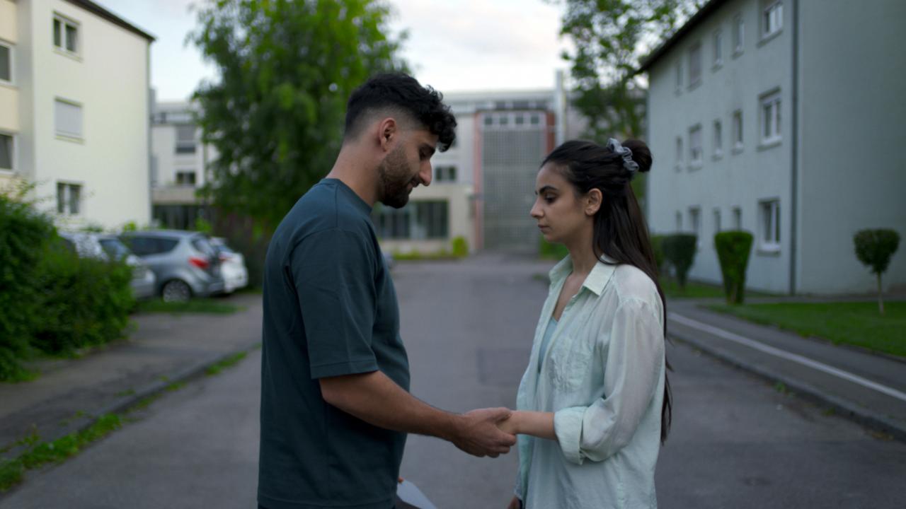 Still aus dem Drama ELAHA: Ein junger Mann und eine junge Frau stehen in einer Wohnstraße, schauen sich an und halten sich an den Händen.