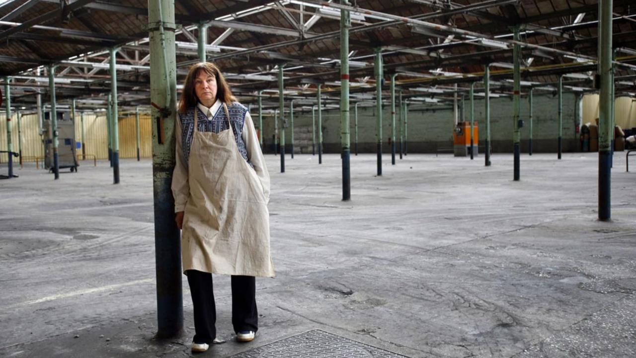 Still aus dem Spielfilm LOUISE HIRES A CONTRACT KILLER: Eine Frau mit weißer Schürze steht in einer leeren Fabrikhalle und lehnt sich an einer der vielen olivgrünen Stahlsäulen an.