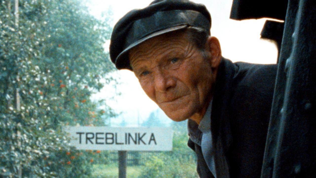 Filmstill aus dem Dokumentarfilm SHOAH: Ein Lokführer schaut aus einer Lok den Betrachter an. Im Hintergrund ist das Ortsschild von Treblinka zu sehen.