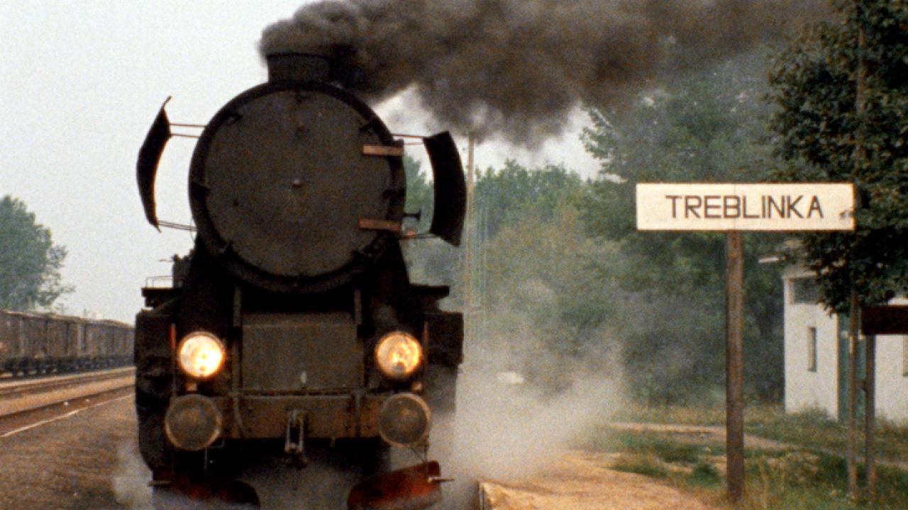 Filmstill aus dem Dokumentarfilm SHOAH: eine Dampflok von vorne, danaben steht ein Schild, auf dem Treblinka steht.