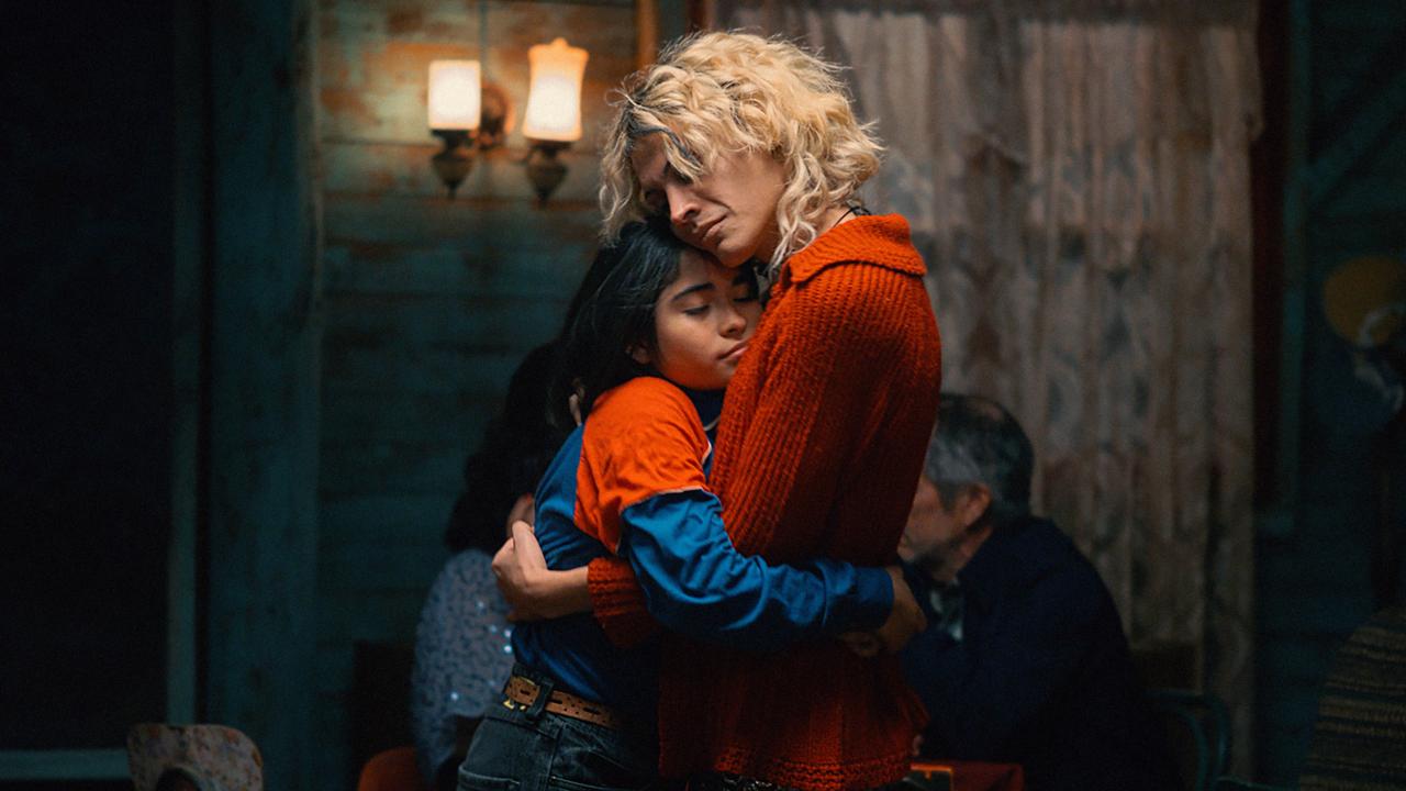 Filmstill aus dem Drama DER GEHEIMNISVOLLE BLICK DES FLAMINGOS: Ein schwarzhaariges Mädchen umarmt eine blonde Person, die eine rote Jacke trägt. Beide haben die Augen geschlossen und stehen in einem spärlich beleuchteteten Raum.