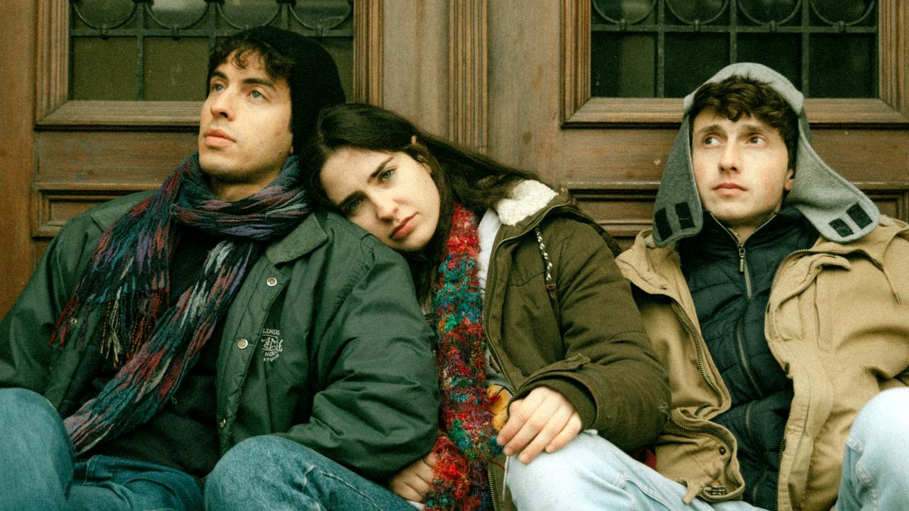 Filmstill aus DELEGATION: Eine junge Frau sitzt zwischen zwei Männern im gleichen Alter vor einer Tür. Sie sind warm angezogen. Die Frau kuschelt sich an einen der Männer an. Alle sehen ersnt aus.