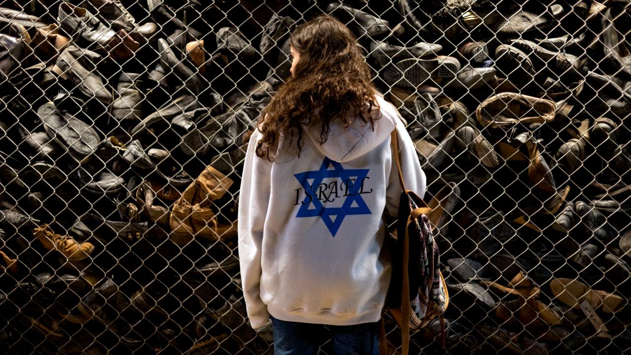 Filmstill aus DELEGATION: Ein Mädchen mit langen Haaren steht mit dem Rücken zur Kamera in der Gedenkstätte Auschwitz vor einer Sammlung von Schuhen. Das Mädchen trägt einen weißen Kapuzenpulli, der mit einem blauen Davidstern und dem Wort Israel bedruckt ist.