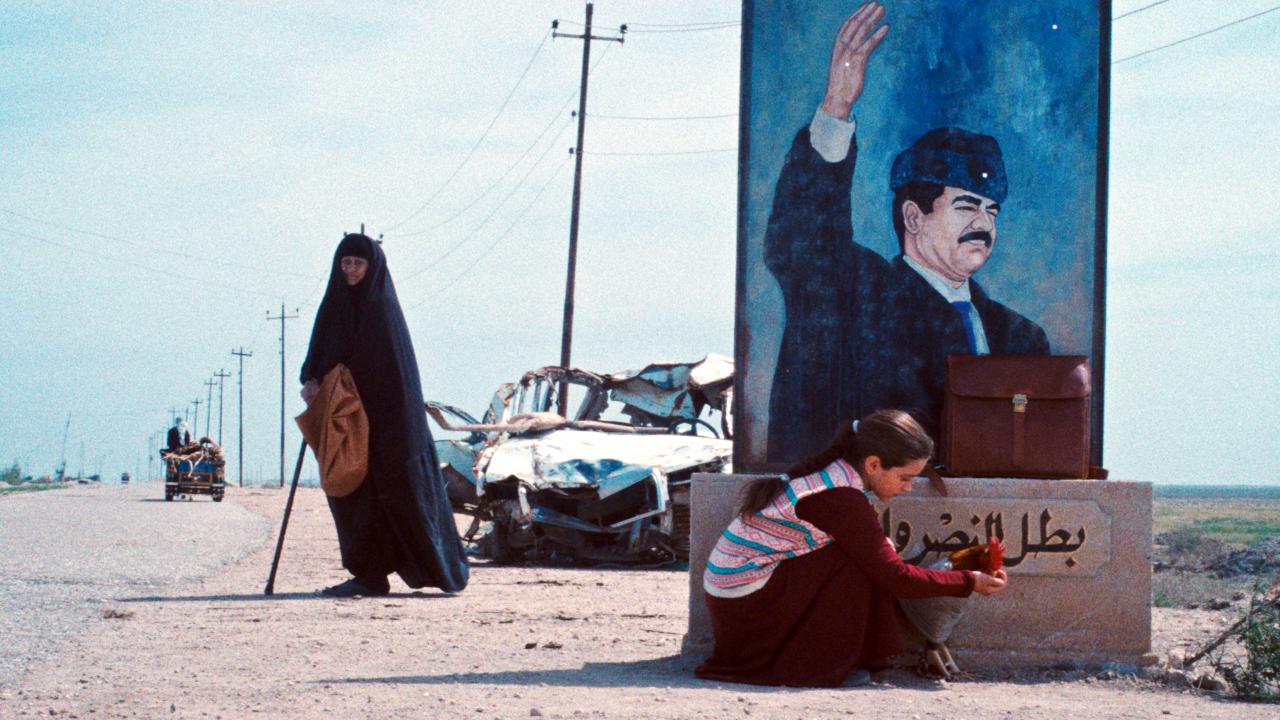 Filmstill aus EIN KUCHEN FÜR DEN PRÄSIDENTEN: Eine alte Frau mit Gehstock steht an einer Straße. Rechts neben ihr hockt ein Mädchen mit einem Hahn vor einem Propagandabild, auf dem der irakische Diktator Saddam Hussein zu sehen ist.