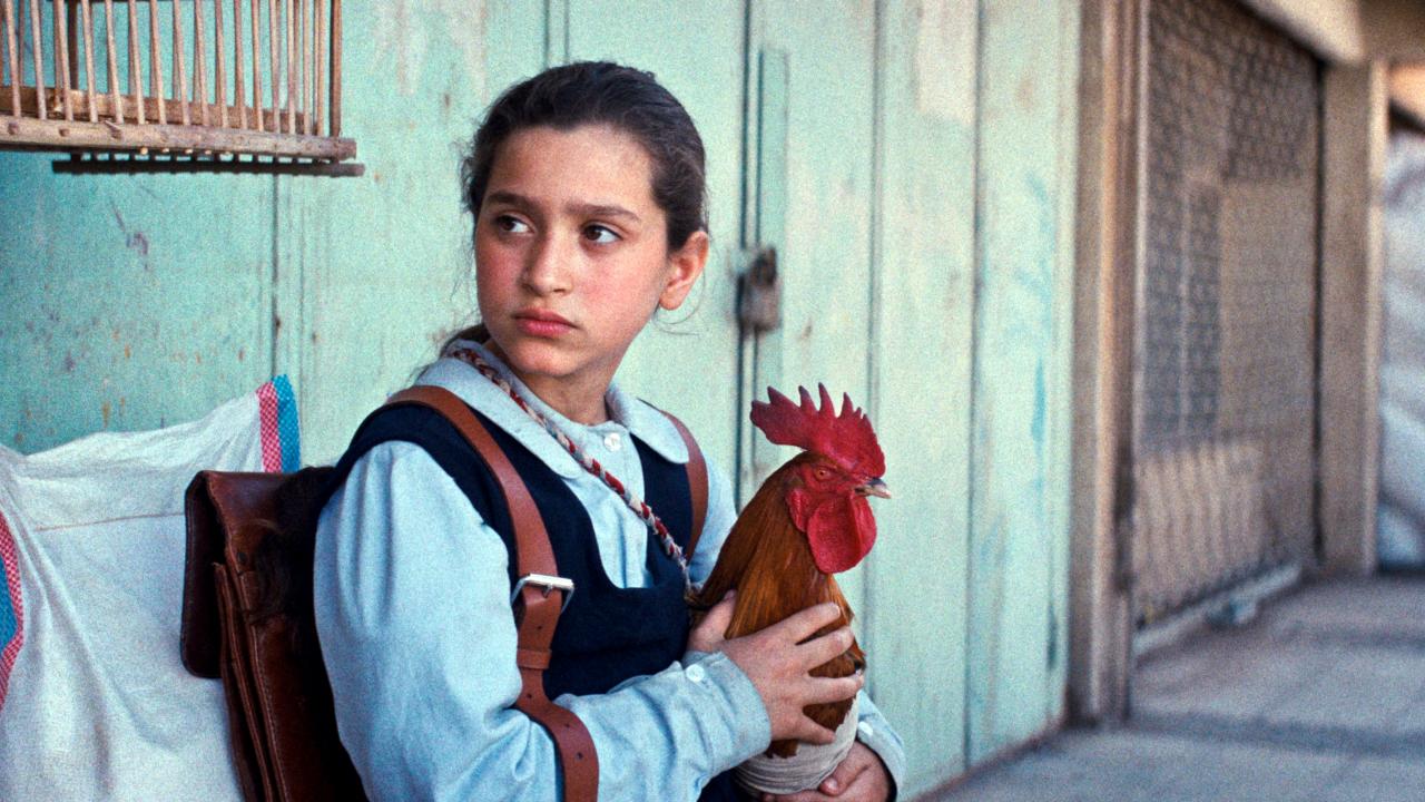 Filmstill aus EIN KUCHEN FÜR DEN PRÄSIDENTEN: Ein Mädchen mit Schulranzen steht an einer Hauswand uns hält einen Hahn in ihren Händen. Es schaut zur Seite.