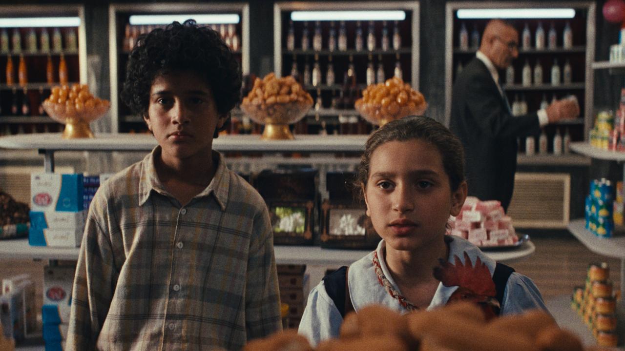 Filmstill aus EIN KUCHEN FÜR DEN PRÄSIDENTEN: Zwei Kinder - ein Junge und Mädchen – stehen an einer Theke in einem Lebensmittelgeschäft. Sie schauen direkt in die Kamera.
