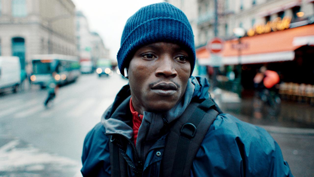 Filmstill aus dem Drama SOULEYMANS GESCHICHTE: Nahaufnahme eines jungen Schwarzen Mannes, der in Winterkleidung gekleidet ist. Er steht auf einer Straßenkreuzung in Paris und schaut ernst.