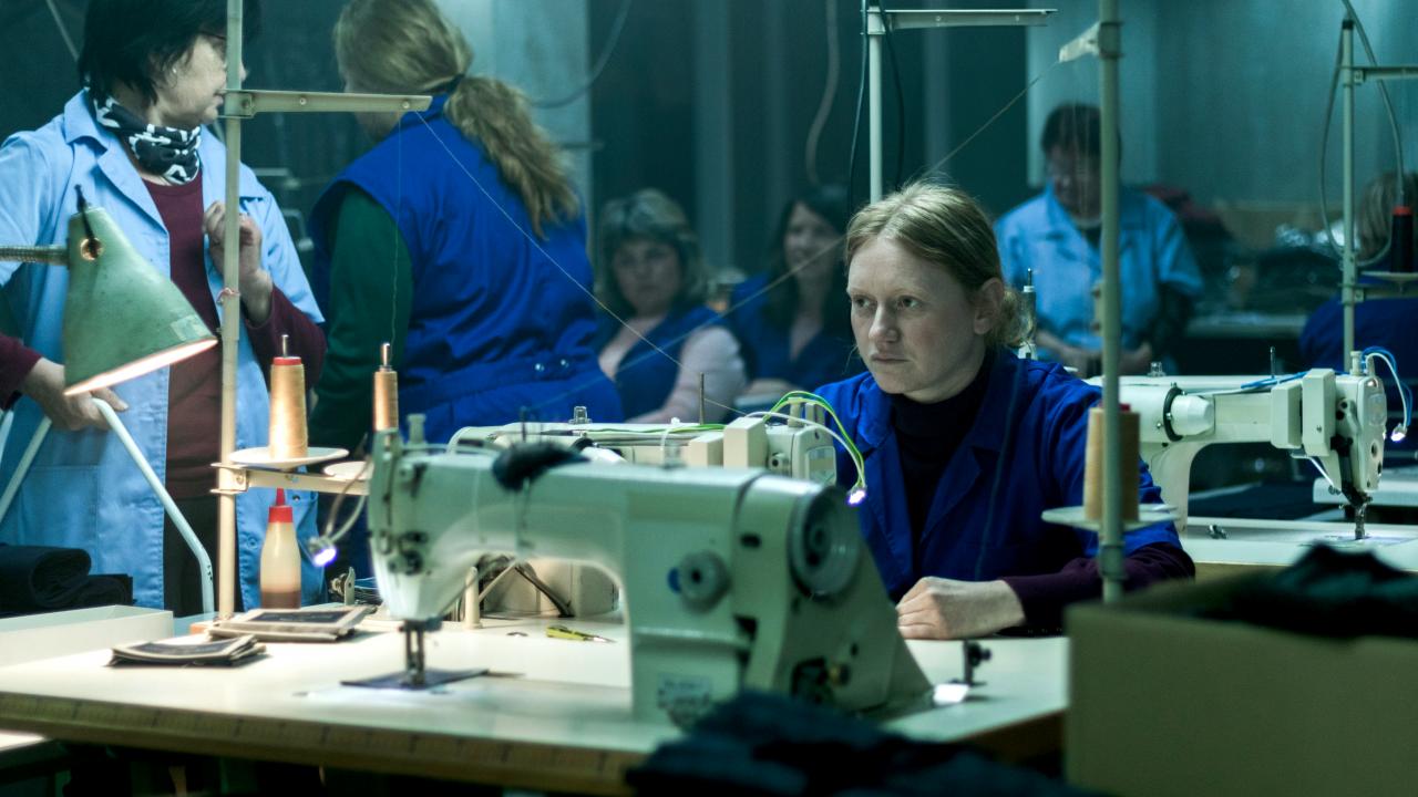 Still aus dem Drama MADE IN EU: In einem fahl beleuchteten Farbrikraum sitzt eine Näherin an ihrer Maschine. Um sie herum sind andere Frauen in Kitteln zu sehen.