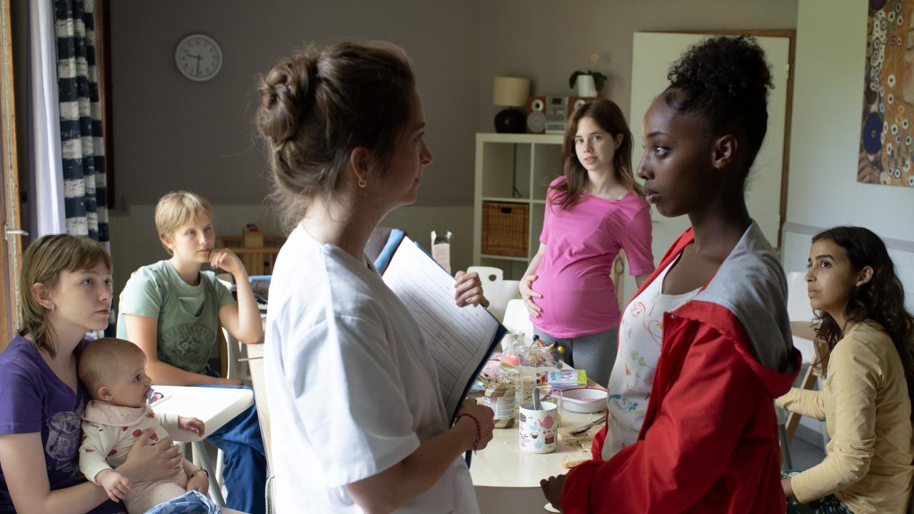 Still aus dem Drama JEUNES MÈRES – JUNGE MÜTTER: Fünf Mädchen im Teenageralter sitzen in einem Raum. Eine ist hochschwanger, eine andere hält ein Baby im Arm. Im Vordergrund spricht ein Mädchen mit einer Krankenschwester.
