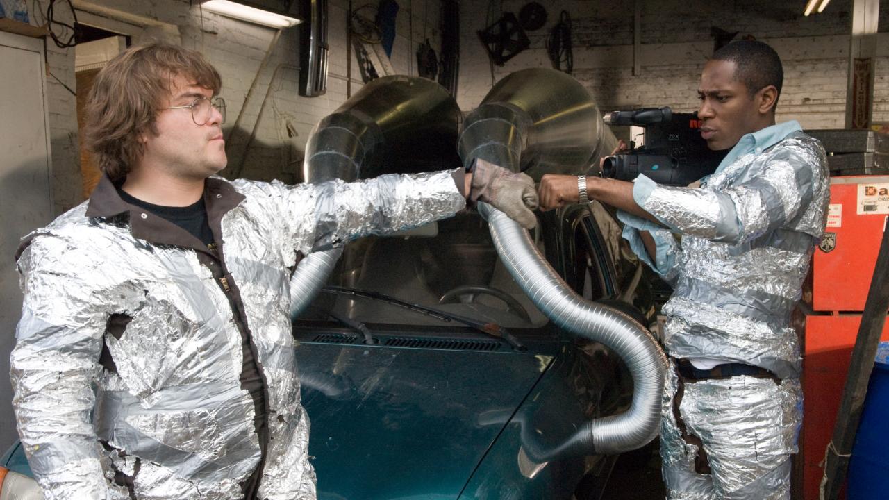 Still aus der Komödie ABGEDREHT: Zwei junge Männer, deren Kleidung mit silberner Folie umwickelt ist, stehen in einer Garage vor einem Auto mit grotesken futuristischen Auspuffrohren versehen ist und stoßen sich die Fäuste an. Der eine hält in seiner anderen Hand eine Videokamera.