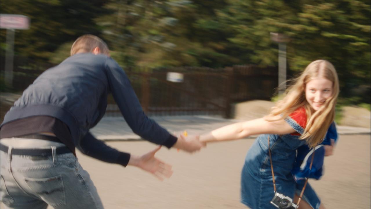 Filmstill aus dem Drama ALS WÄRE ES LEICHT: Eine junge Frau mit langen blonden Haaren zieht lächelt einen Mann hinter sich her. Sie scheinen Rollschuhe zu laufen oder zu rennen. Das Bild ist dynamisch..