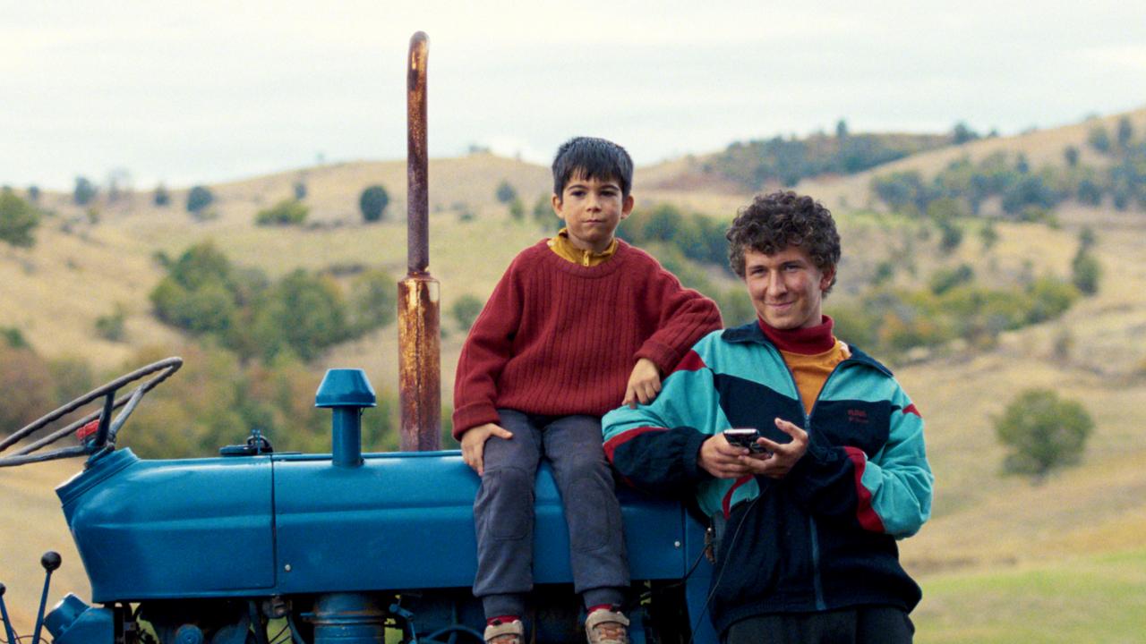 Still aus dem Drama DJ AHMET: Vor einer weiten Hügellandschaft steht ein blauer, alter Traktor. Auf ihm sitzt ein kleier Junge und schaut direkt in die Kamera. Neben ihm steht ein etwa 15-Jähriger und lächelt.