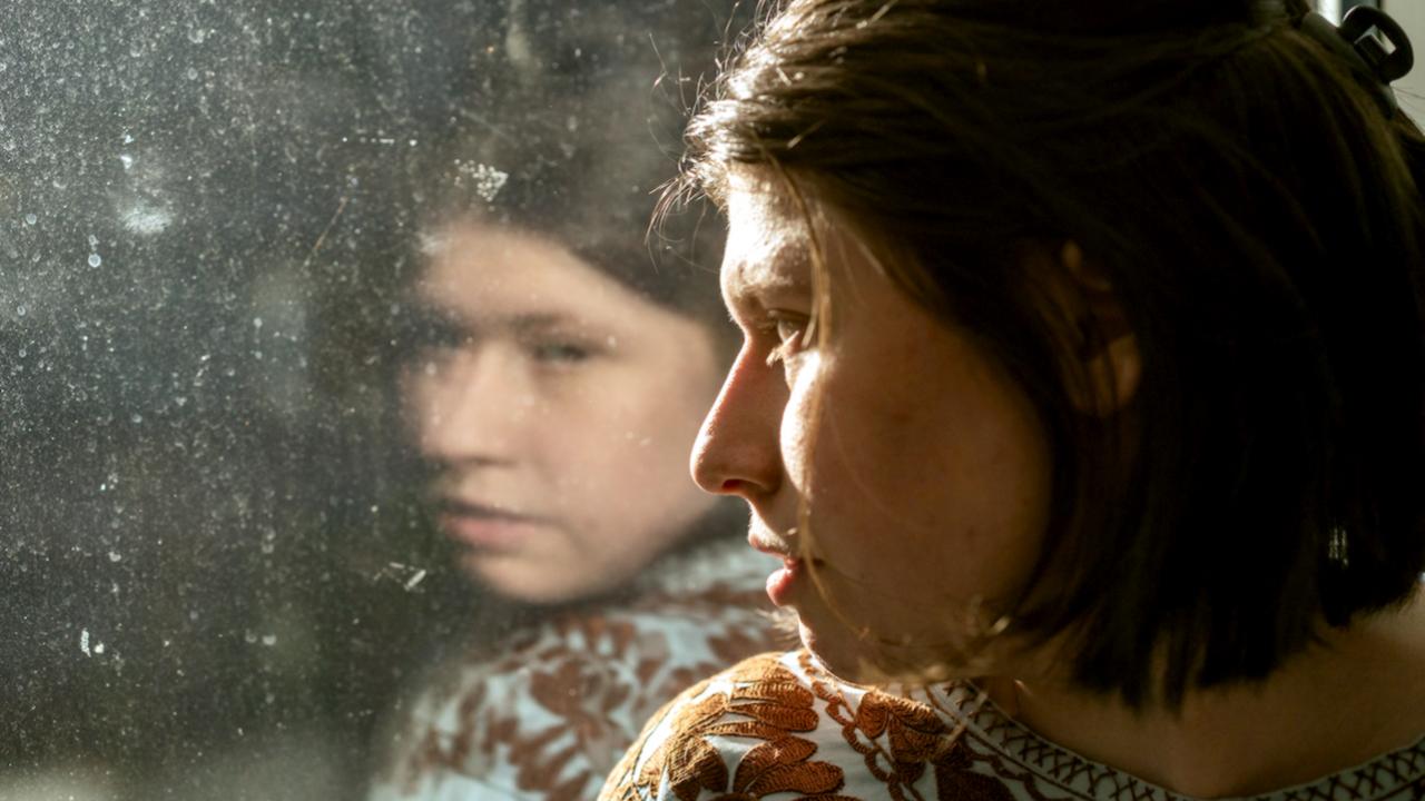 Still aus dem Drama LUISA. Eine junge Frau sitzt vor einem Fenster und schaut nach draußen. Sie ist eher im Profil zu sehen. In der Schreibe ist ihr Spiegelbild zu sehen.