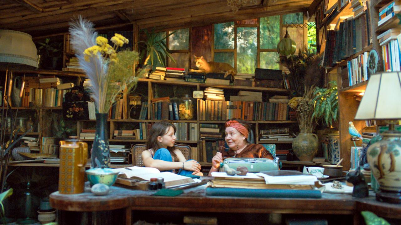 Still aus dem Drama I ACCIDENTLY WROTE A BOOK: Ein Mädchen im Teenageralter und eine ältere Frau sitzen vor einem Tisch und unterhalten sich. Das Zimmer ist voll mit Büchern.