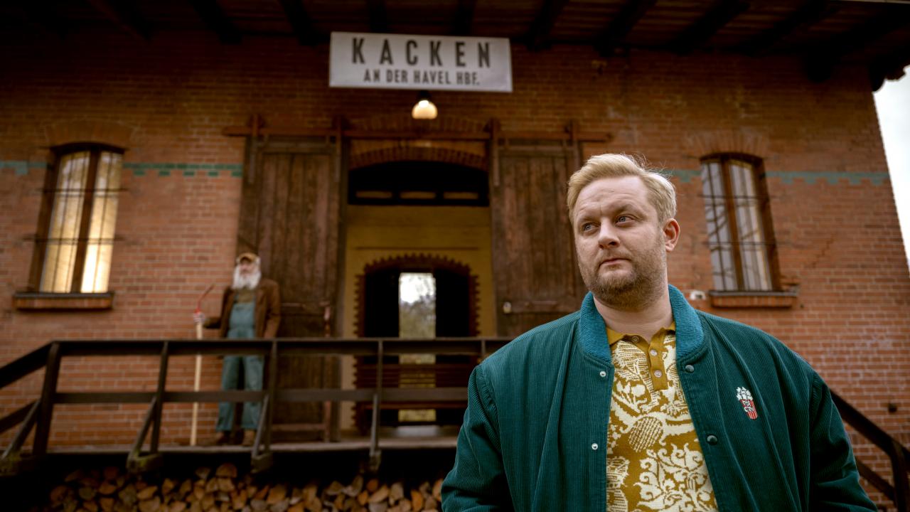 Still aus der Serie KACKEN AN DER HAVEL: Ein Mann um die 30 steht vor einem Backsteingebäude, an dem ein Ortsschild mit der Aufschrift "Kacken an derr Havel" angebracht ist. Das Bild ist leicht von unten aufgenommen.